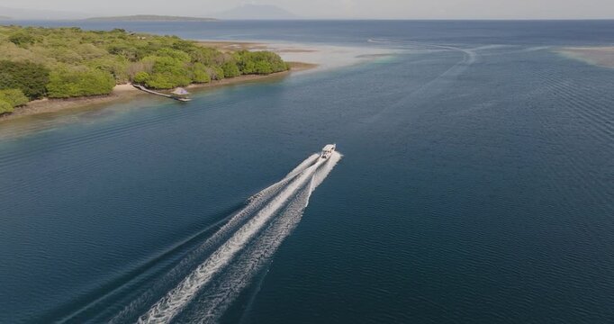 Aerial drone shot following a small boat leaving the calm lagoon of West Bali National Park, Indonesia, with rippling wake patterns spreading across deep blue tropical waters near mangrove coastline