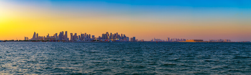 Wide panoramic view of Doha skyline in Qatar photographed at sunset with high-rise buildings silhouetted above the Gulf waters