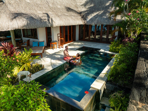 Aerial view of a couple enjoying a floating breakfast in a serene pool surrounded by lush greenery and a thatched-roof villa, Prince Maurice, Flacq District, Mauritius.