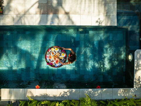 Aerial view of a person with a floating tray of food in a pool, framed by the geometric precision of white tiles and verdant foliage, Prince Maurice, Flacq District, Mauritius.