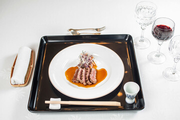 A modernly arranged dish with thinly sliced ​​meat in an orange sauce, served on a white plate with accompanying chopsticks.