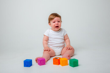 a baby boy in a white bodysuit is sitting on a white background playing with blocks and crying very upset