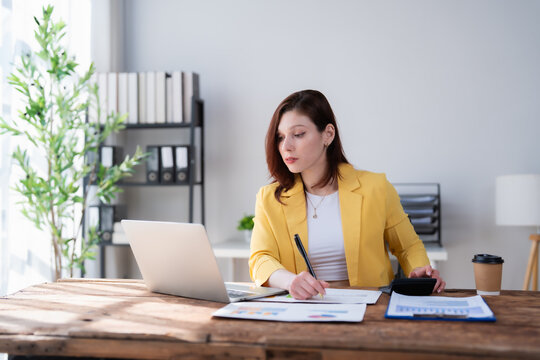 Young woman working on financial planning and paperwork at a modern office desk, calculating numbers with a calculator and referencing data on her laptop