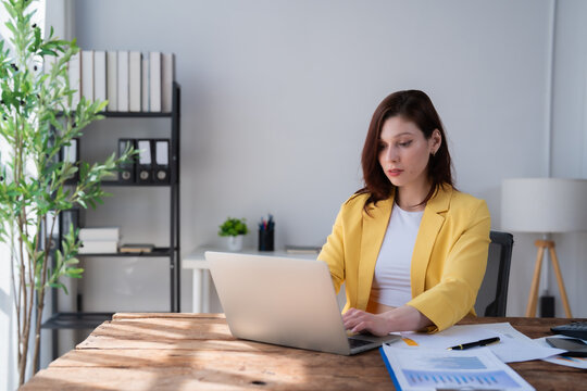 Woman in a yellow blazer focusing on her laptop, analyzing financial documents and charts, symbolizing business growth, freelance work, and digital entrepreneurship in a modern office setup