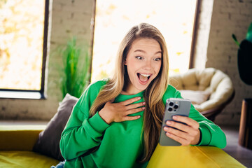 Naklejka premium Joyful young woman in green sweatshirt reacting to exciting news with her smartphone in a cozy interior loft