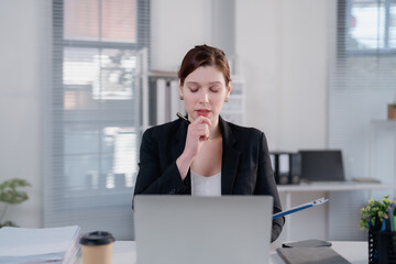Young businesswoman contemplating ideas and working on her laptop at an office desk, holding a pen...
