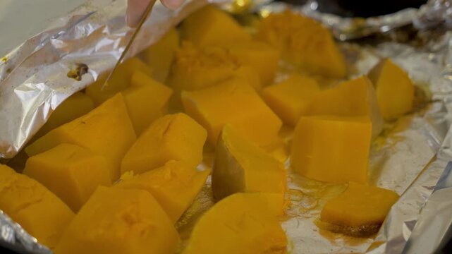 Knife pierces roasted pumpkin to test softness. Female hand opens foil in tray. Autumn harvest meal prepared for Halloween. Dish reflects warmth, care, and seasonal tradition