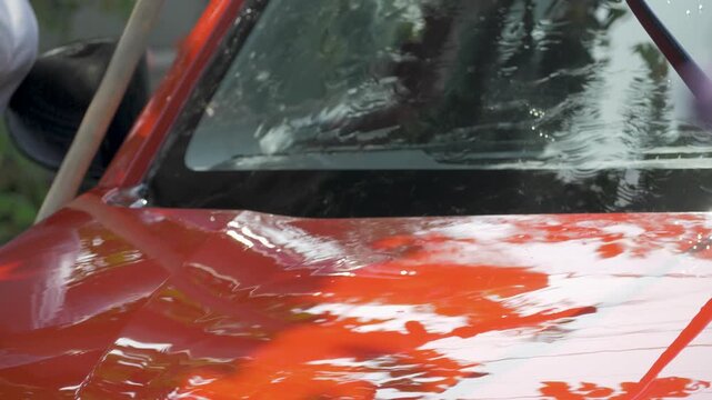 Red car under water stream. Female driver hand removes dirt from surface with yellow sponge. Clean, polished finish achieved with care. Represents dedication to maintaining vehicle shine