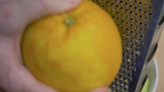Housewife hand moves fruit across grater surface into fine pieces. Orange peel becomes zest for muffin recipe. Preparation and purpose in kitchen routine. Shredder kitchen utensil used to shred foods