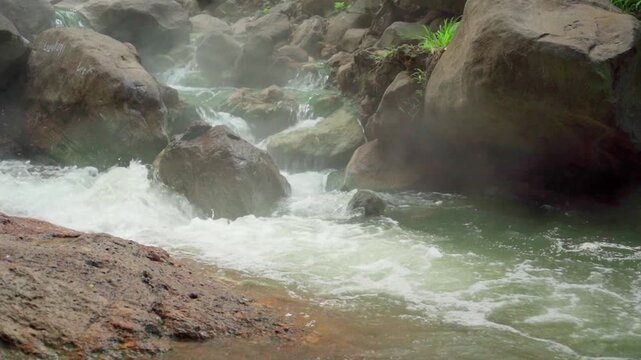 Fast flowing tropical stream rushing between large rocks near Malanage Hot Spring in Indonesia, highlighting natural spring water and jungle landscape.