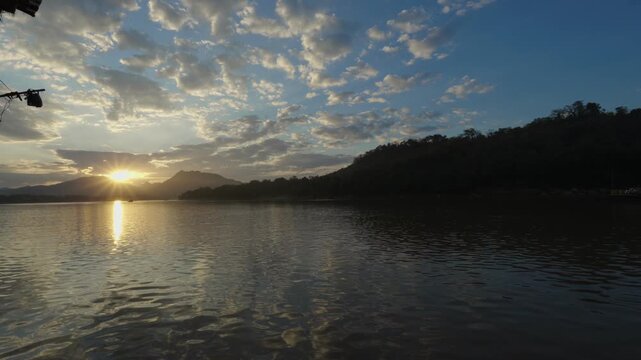 View of the setting Sun behind the mountais at the Mekong River with light cloudy sky and calm water in the foreground, with mountain silouettes in the background.