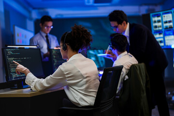 Obraz premium Friendly female IT support agent wearing headset working on computer in dark blue office. Professional customer service operator helping client in night shift control center room.