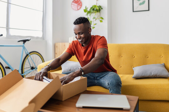Happy African man opening delivery box with new clothes purchased online at home. Black male customer receiving cardboard parcel box. Online shopping and home delivery concept