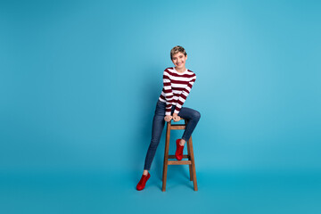 Fototapeta premium Young woman sits on a wooden stool against a bright blue background wearing a striped sweater jeans and red shoes posing for a stylish fashion lifestyle shoot
