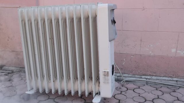 Defective white electric oil heater starting to smoke from the bottom, indicating a dangerous malfunction, short circuit, or potential fire hazard due to overheating electrical components