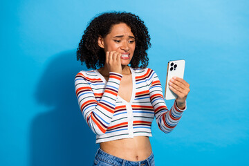 Young chic woman in striped cardigan on blue background looks at phone with worried expression