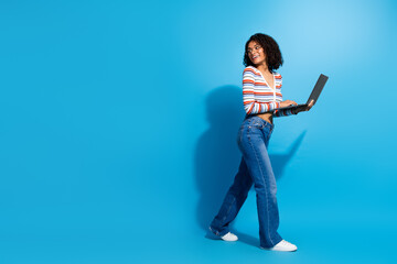 Young chic woman with laptop walks against blue backdrop smiling as she works remotely and...