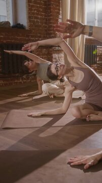 Full vertical shot of three Caucasian girls sitting with crossed legs on mats, performing alternating side stretch with arm extended over head, while practicing yoga or pilates in studio