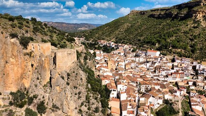 Aerial drone view of Chulilla in Spain showing a mountain village nestled between dramatic limestone canyon cliffs. Detailed landscape of terracotta rooftops, river valley and surrounding mountains.