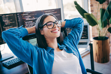 Happy female programmer in a modern loft office coding at desk with multiple monitors and a relaxed...