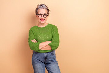 Mature woman with short grey hair wearing a green knit sweater stands with arms crossed and smiles warmly against a beige studio background © deagreez
