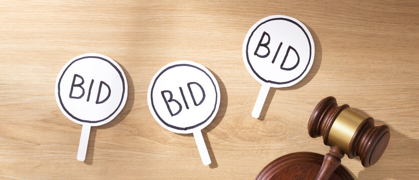 Wooden gavel on sound block next to multiple white "BID" signs on a wooden desk, symbolizing auction bidding and competitive sales. Panoramic top view.