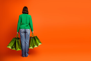 Fototapeta premium Back view of a young chic woman in a green sweater carrying multiple green shopping bags against a bright orange background