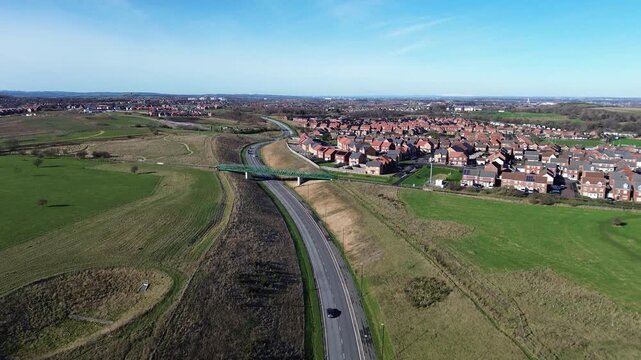 Drone flyover of St Benedict's Way, Ryhope, gliding above road, bridge and new housing estate beside open green fields under clear blue sky in Sunderland, North East England.