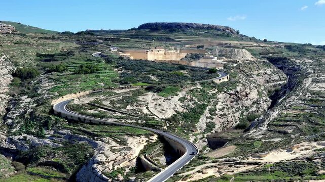 High angle wide shot of winding asphalt road snaking through sun-drenched terraced hills of Gozo Malta under clear blue sky during lush spring season featuring rugged limestone cliffs and greenery