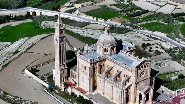 Aerial view of Ta Pinu Basilica in Gozo Malta during a bright spring day featuring the iconic limestone architecture against lush green fields and terraced landscapes under a clear blue sunny sky