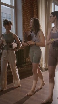 Full vertical tracking shot of three sporty young Caucasian female friends with yoga mats walking and chatting in corridor of loft yoga studio to group class