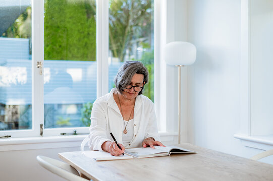 A woman sits at a table, writing in a journal in a well-lit room with windows