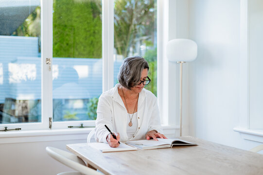 Senior woman sits at a table reading and writing in a book