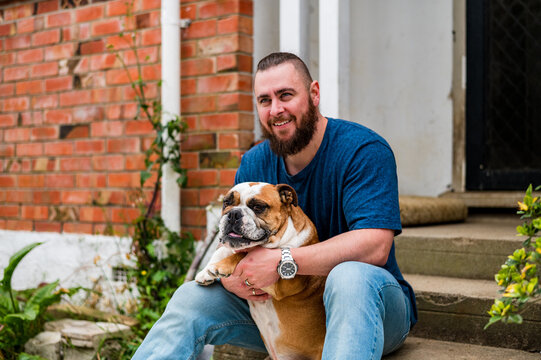 Man smiles while holding bulldog on steps outside brick house in New Zealand.