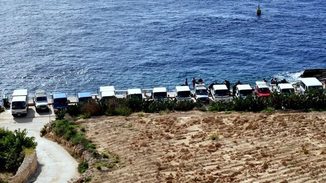 Rugged white offroad vehicles parked along the rocky coastline of Gozo Malta under a bright summer sun as turquoise Mediterranean waves crash against the shore captured from a high angle view