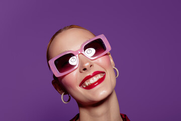 Smiling woman with bright red lipstick wears large pink sunglasses and hoop earrings against purple background. Concept of fashion, beauty, and confident style portrait. © SHOTPRIME STUDIO