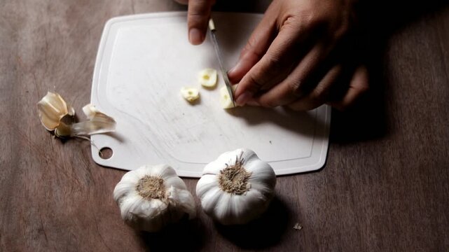 Slicing garlic cloves on a plastic cutting board using a kitchen knife, showing food preparation activity.