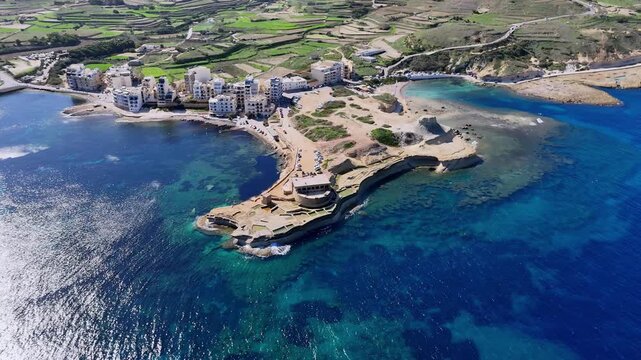 Aerial drone view of Xlendi Bay Gozo Malta featuring crystal clear turquoise Mediterranean waters limestone cliffs white coastal buildings lush green terraced fields and sunny spring weather