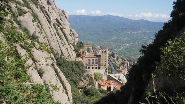 A high-vantage perspective of the Santa Maria de Montserrat Abbey nestled against the serrated mountain spires. Captures the monastery's architecture integrated into the sheer rock walls.