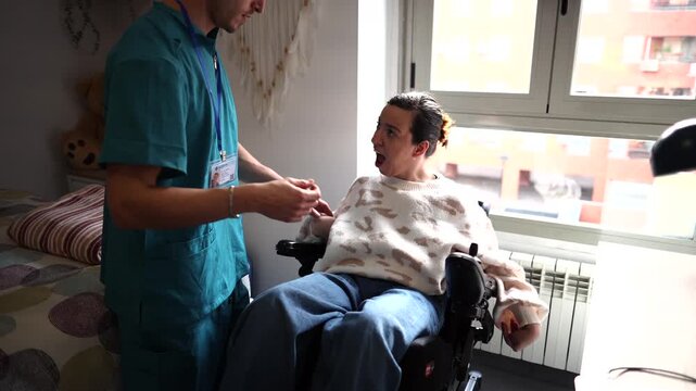 Young caregiver helping a smiling woman with a disability, assisting her to sit comfortably in her electric wheelchair in a sunlit room, demonstrating professional healthcare support and assistance