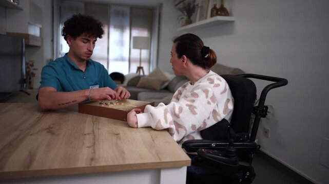 Male nurse enjoying a game of backgammon with a disabled woman in a wheelchair, providing companionship and support during a home visit as part of her rehabilitation and healthcare routine