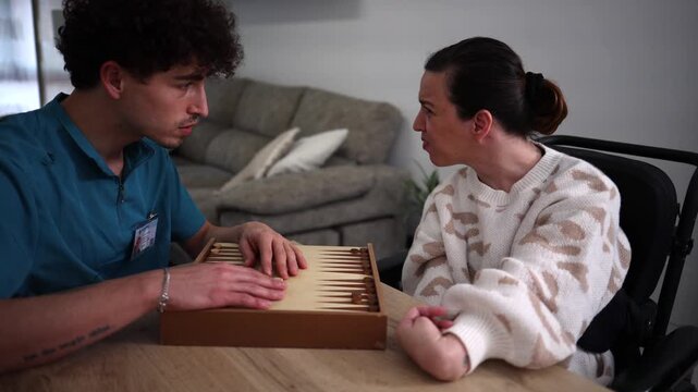 Male caregiver providing companionship and cognitive stimulation to a disabled woman in a wheelchair by playing a game of backgammon together at a table in a care facility