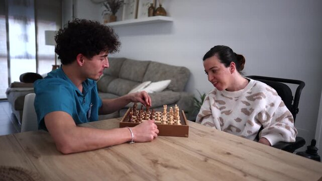 Cheerful disabled woman in a wheelchair having fun and laughing while playing a game of chess with her male caregiver at home, promoting inclusive leisure activities and positive patient care