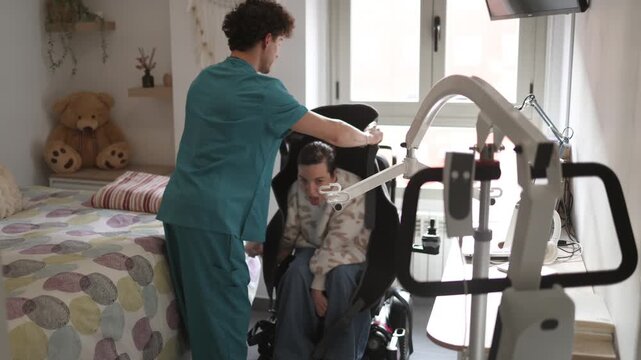 Caring male nurse placing a hoist sling behind a young woman with a physical disability sitting in a wheelchair, preparing her for transfer with a patient lift in her bedroom