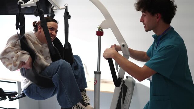 Caring male nurse operating a patient lift to carefully lower a disabled woman in a rehabilitation center, providing essential support and mobility assistance during a therapy session