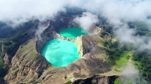 Drone aerial of Kelimutu volcano featuring its iconic turquoise crater lakes surrounded by dramatic volcanic terrain and drifting clouds in Flores, Indonesia.