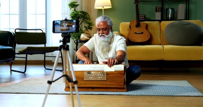 Indian mid age bearded man recording harmonium playing session on smartphone for online class at home, stylish mature music tutor creating classical lesson video with tripod setup in modern studio