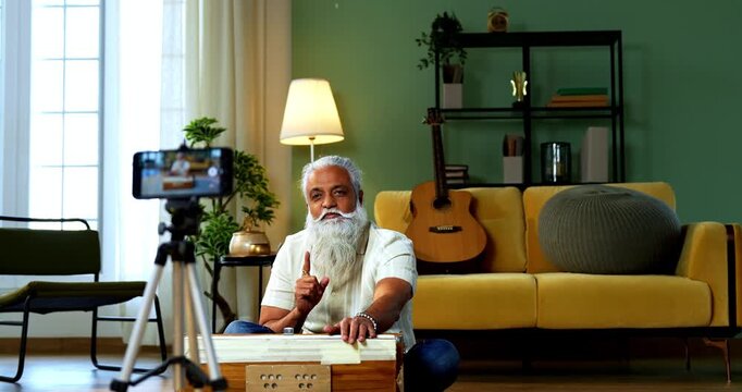 Indian mid age bearded man recording vocals music session on smartphone for online class at home, stylish mature tutor teaching Indian classical singing with harmonium in modern studio