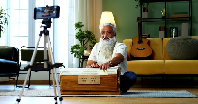 Indian mid age bearded man recording vocals music session on smartphone for online class at home, stylish mature tutor teaching Indian classical singing with harmonium in modern studio