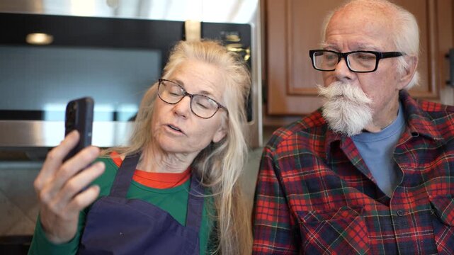 Elderly man and his mature daughter talk to a family member through video chat in their kitchen during the afternoon.